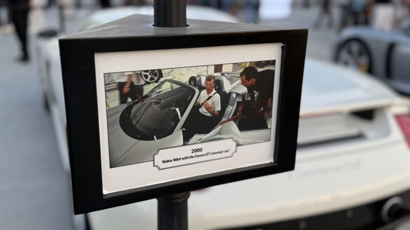 A framed photo on display shows two people beside a silver convertible car, with a caption reading, "2000. Walter Röhrl with the Carrera GT concept car." Feature Exclusive: 2025 Icons of Porsche Dubai.
