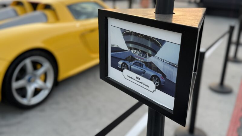 A yellow sports car is parked next to a sign showing a photo and details of a 2004 Porsche Carrera GT, displayed outdoors with a barrier in the background—Feature Exclusive: 2025 Icons of Porsche Dubai.
