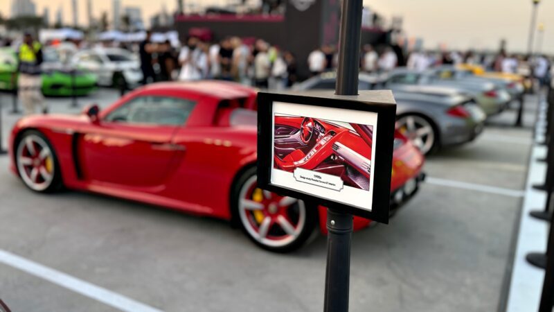 A red sports car is parked at an outdoor car show, with a display sign featuring an interior photo and description of the vehicle in the foreground. Feature Exclusive: 2025 Icons of Porsche Dubai can be seen in the background among other cars and people.