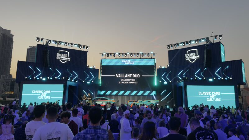 A large crowd gathers in front of a brightly lit outdoor stage featuring classic cars and digital screens at sunset—Feature Exclusive: 2025 Icons of Porsche Dubai.