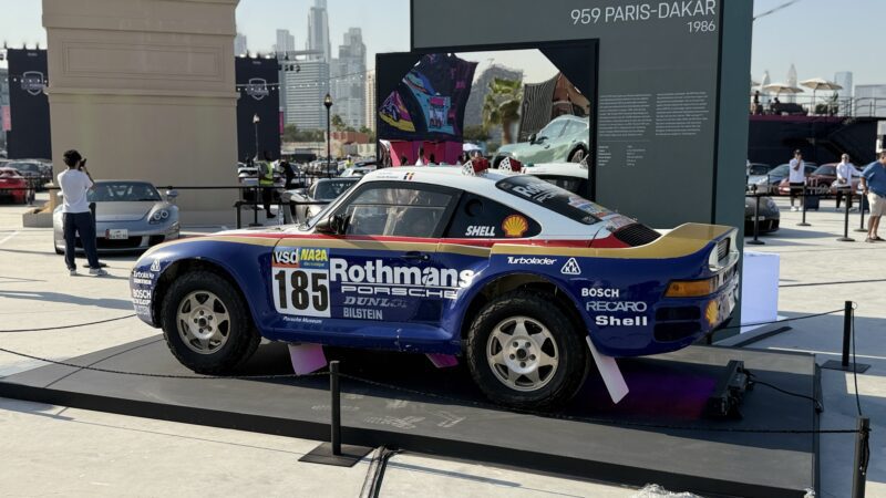 Feature Exclusive: 2025 Icons of Porsche Dubai – A classic Rothmans-liveried Porsche 959 Paris-Dakar rally car on outdoor display, with informational signage and city buildings visible in the background.