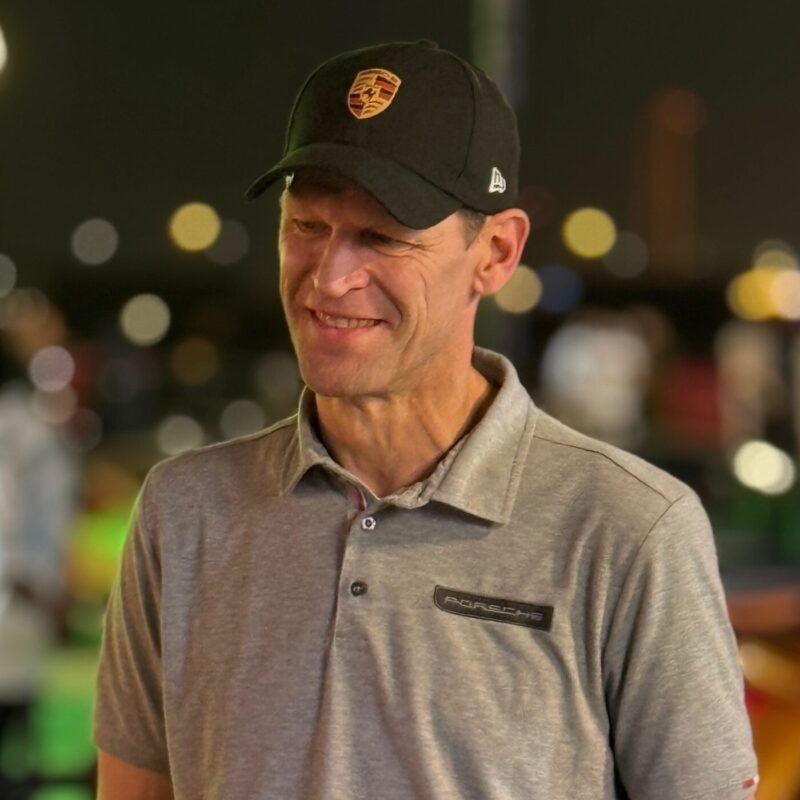 A man wearing a black Porsche cap and gray Porsche-branded polo shirt stands outdoors at night with blurred lights in the background, featured exclusively at the 2025 Icons of Porsche Dubai.