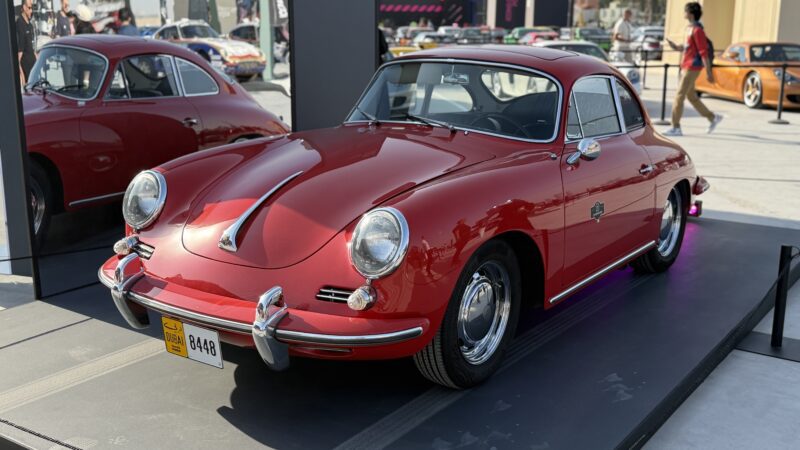 A classic red Porsche coupe is displayed on a platform outdoors, its side reflected in a mirror. Feature Exclusive: 2025 Icons of Porsche Dubai showcases this and other vintage cars in the background.
