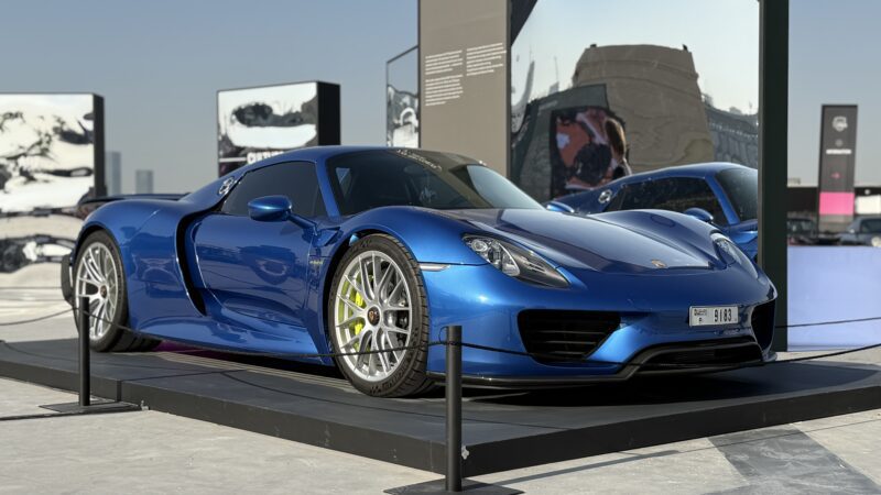 A blue Porsche 918 Spyder sports car is displayed on a platform outdoors, surrounded by a barrier, with modern buildings and artwork in the background. Feature Exclusive: 2025 Icons of Porsche Dubai.