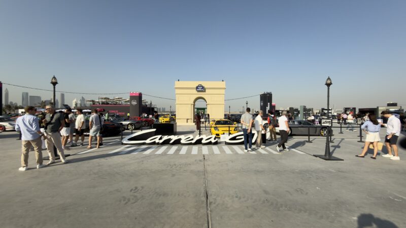 People walk around an outdoor car exhibition featuring luxury sports cars, with a large "Carrera GT" sign on the ground and a small building structure in the center. Feature Exclusive: 2025 Icons of Porsche Dubai.