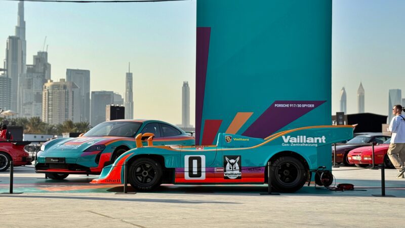 Feature Exclusive: 2025 Icons of Porsche Dubai—a teal and orange vintage race car with number 0 is displayed outdoors among modern sports cars, set against a city skyline and a tall sign reading "Porsche 911 GT3 GPRR.