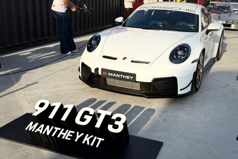 A white Porsche911 GT3 992.2 with a Manthey Kit is showcased outdoors; a black sign in front reads "911 GT3 MANTHEY KIT." This setup delivers 1190 pounds of downforce and achieved a 6:52 Nürburgring lap.