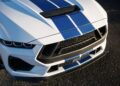 Close-up of the front end of a white sports car with blue racing stripes, showcasing the grille, headlights, and hood scoop on a paved surface. Review: 2025 Shelby GT350 highlights its bold design details.