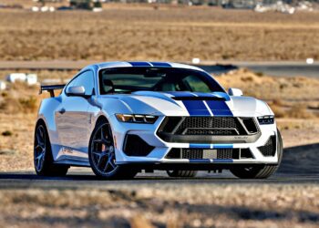 A white sports car with blue racing stripes, the Review: 2025 Shelby GT350, is parked on a paved road in a desert landscape with dry grass and distant buildings.