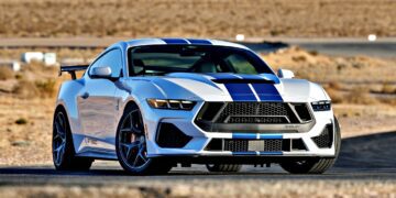 A white sports car with blue racing stripes, the Review: 2025 Shelby GT350, is parked on a paved road in a desert landscape with dry grass and distant buildings.