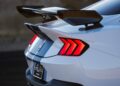 Close-up of the rear of a white sports car with red taillights, black racing stripes, and a large carbon fiber spoiler. Review: 2025 Shelby GT350 highlights its bold design and performance-driven features.