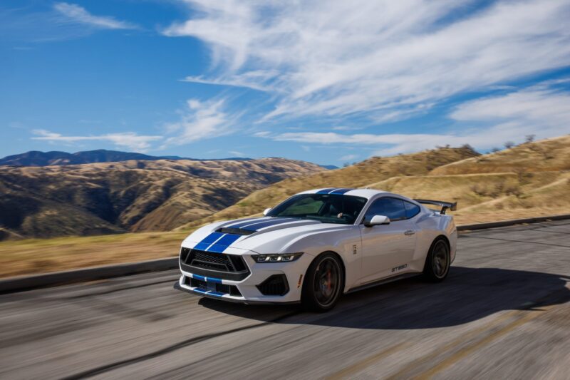 A white sports car with blue racing stripes—like the one featured in our Review: 2025 Shelby GT350—drives on a winding road through a dry, hilly landscape under a blue sky with scattered clouds.