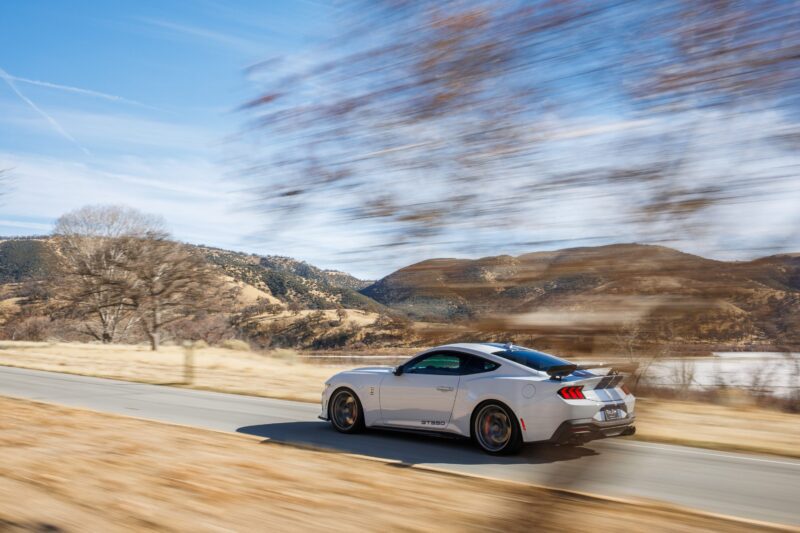 A silver sports car, the 2025 Shelby GT350, drives quickly down a rural road with blurred trees and hills in the background under a blue sky. Review: Experience pure performance with this exhilarating ride.