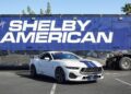 A white Ford Mustang with blue racing stripes, likely the star of a Review: 2025 Shelby GT350, is parked in front of a trailer with large "Shelby American" text.