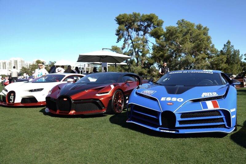 Three Bugatti sports cars are parked on grass at an outdoor auto show, with trees, people, and city buildings visible in the background—capturing the spirit of Wynn Concours 2025: Hypercars World Debuts And Elevated Hospitality.