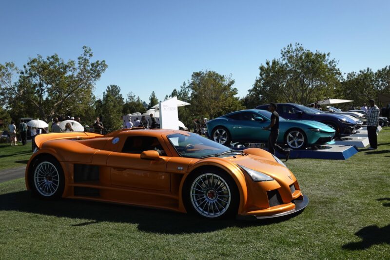 An orange sports car displayed on grass at the Wynn Concours 2025, Las Vegas’ premier automotive event for hypercars world debuts and elevated hospitality, with other cars and spectators visible in the background.