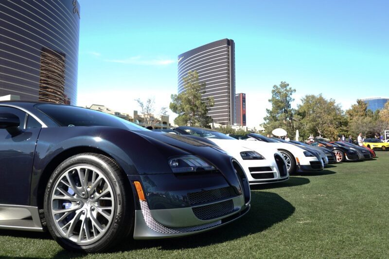 A row of luxury sports cars, including multiple Bugatti models, are parked on a grassy area at the Wynn Concours 2025: Hypercars World Debuts and Elevated Hospitality—Las Vegas’ premier automotive event—set against modern high-rises under a clear blue sky.