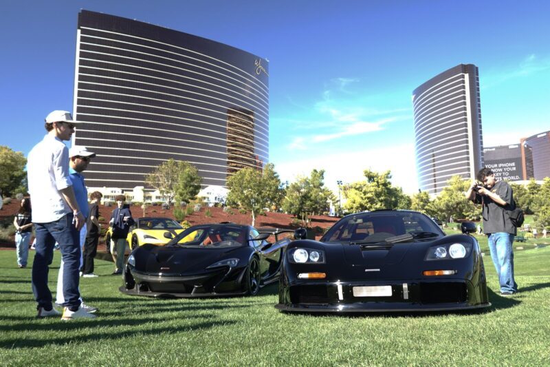 A group of people stand near luxury sports cars displayed on grass at Wynn Concours 2025: Hypercars World Debuts and Elevated Hospitality, set against tall modern buildings and a clear blue sky at Las Vegas’ premier automotive event.