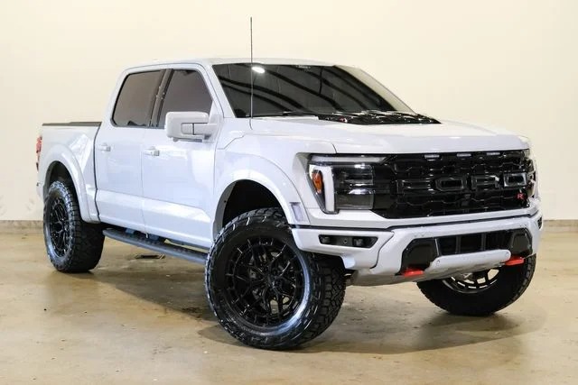 A white Ford F-150 Raptor pickup truck with black wheels and tinted windows showcases style and comfort, parked indoors on a concrete floor.