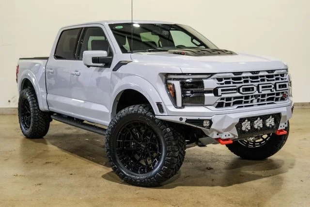 A white Ford F-150 Raptor pickup truck with off-road tires, custom front bumper, and black wheels embodies the comfort and style found in today’s luxury trucks, parked indoors on a concrete floor.