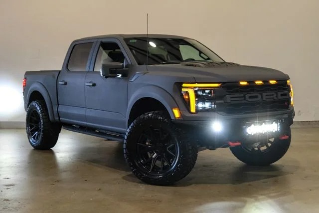 Matte black Ford pickup truck with large off-road tires, illuminated front lights, and amber marker lights parked indoors against a plain background, showcasing bold style and the rugged comfort found in today’s luxury trucks.