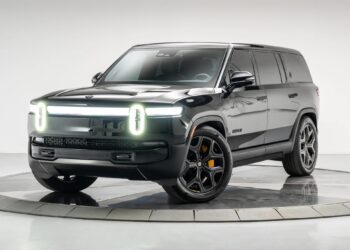 A black Rivian SUV, representing the new wave of affordable luxury SUVs, is parked on a circular platform in a studio setting, featuring distinctive illuminated front lights and black alloy wheels.