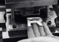 A hand holding a metallic letter "R" near a metal stamping machine in a workshop setting, reminiscent of the precision seen as Range Rover expands its 2026 Sport lineup with SV Black and SV Carbon editions.
