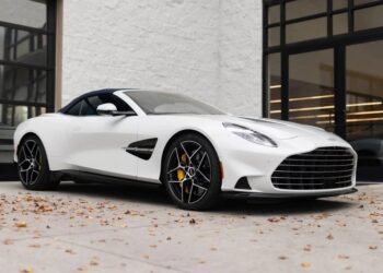 A white Aston Martin luxury car convertible is parked on a concrete surface near a building with glass windows; fallen leaves are scattered on the ground.