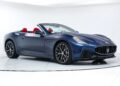 A blue Maserati convertible with red interior seats, this luxury car is displayed in a white showroom with a clear view of the front and side.