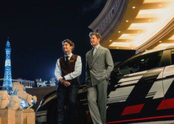 Two men in formal attire stand beside a black car at night in Vegas, with bright lights and a replica of the Eiffel Tower visible in the background.