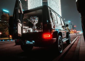 A luxury SUV with open rear doors displays a large reflective trophy in the trunk, parked on a city street at night with illuminated buildings in the background.
