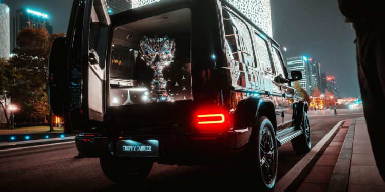 A luxury SUV with open rear doors displays a large reflective trophy in the trunk, parked on a city street at night with illuminated buildings in the background.