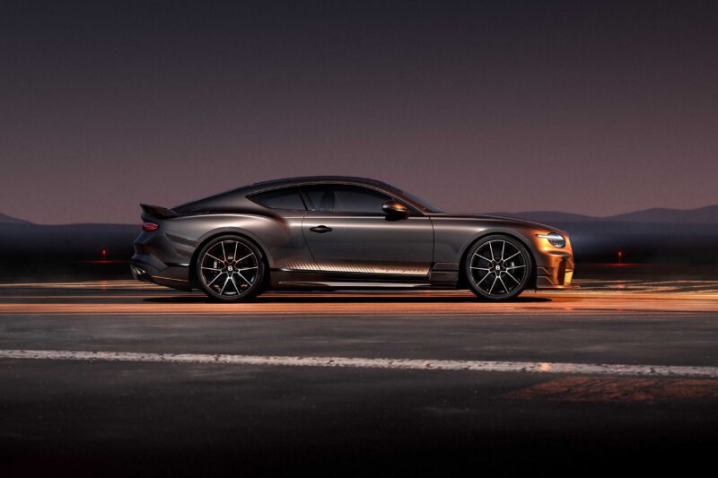 A matte black Bentley Supersports is parked on a road at dusk, viewed from the side, with mountains and a dark sky in the background—accentuating its driver-focused design.