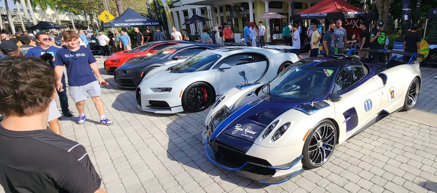 A row of luxury hypercars, including a Pagani, Bugatti, and the Praga Bohema, is displayed at a South Florida Automotive show as people observe and take photos.