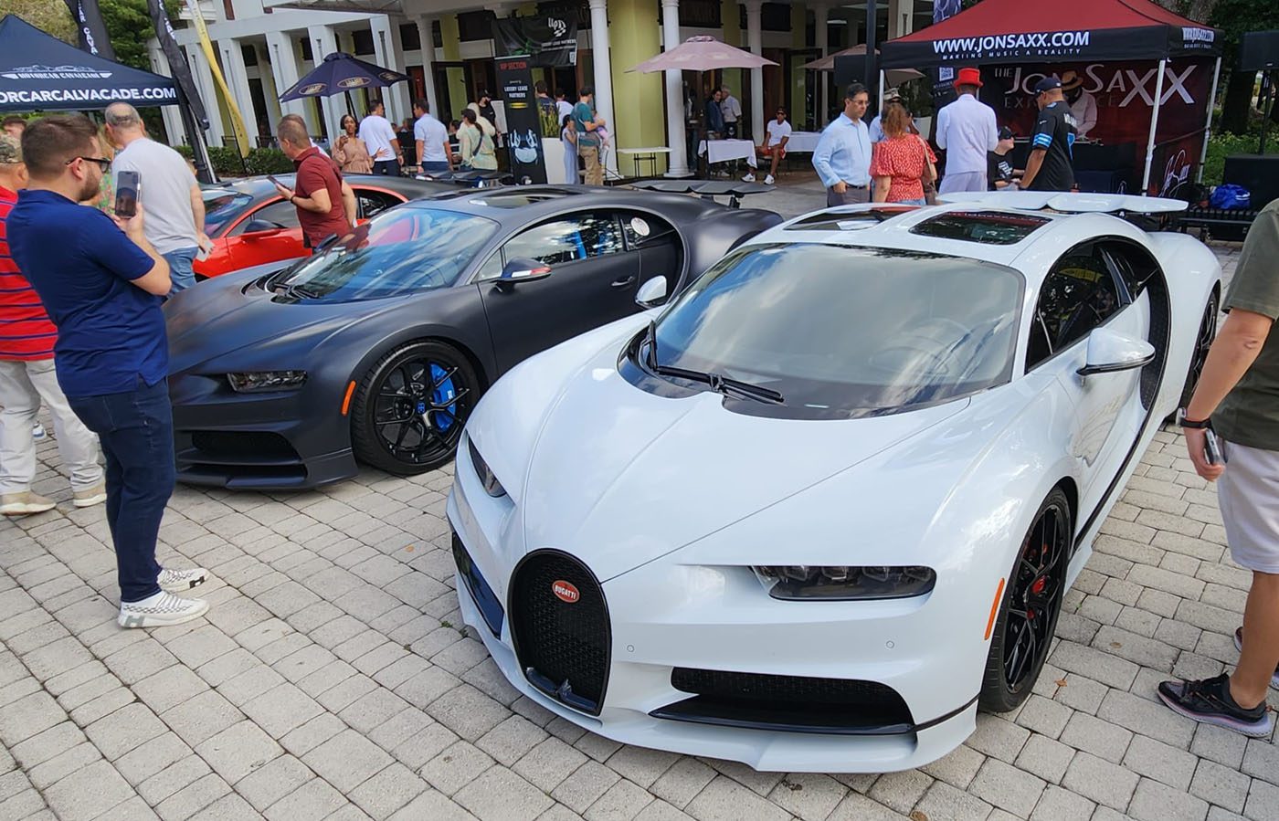 A white Bugatti Chiron and a dark grey Bugatti Chiron, two stunning hypercars, are parked side by side at an outdoor South Florida Automotive car show, surrounded by people taking photos.