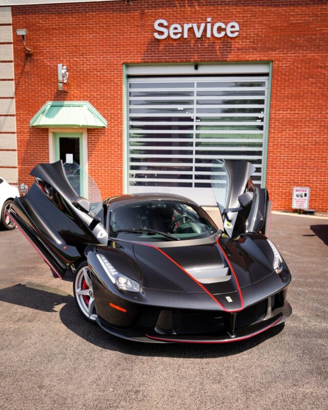 A black sports car with its butterfly doors open is parked in front of a brick building labeled "Service," showcasing Ferrari Lake Forest’s commitment to excellence and its pursuit of Top Dealer Awards.