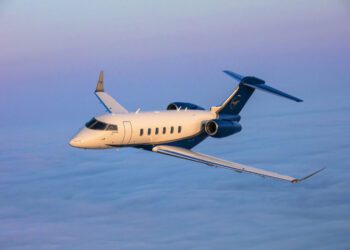 A blue and white Airshare private jet flies above a blanket of clouds during sunset or sunrise.