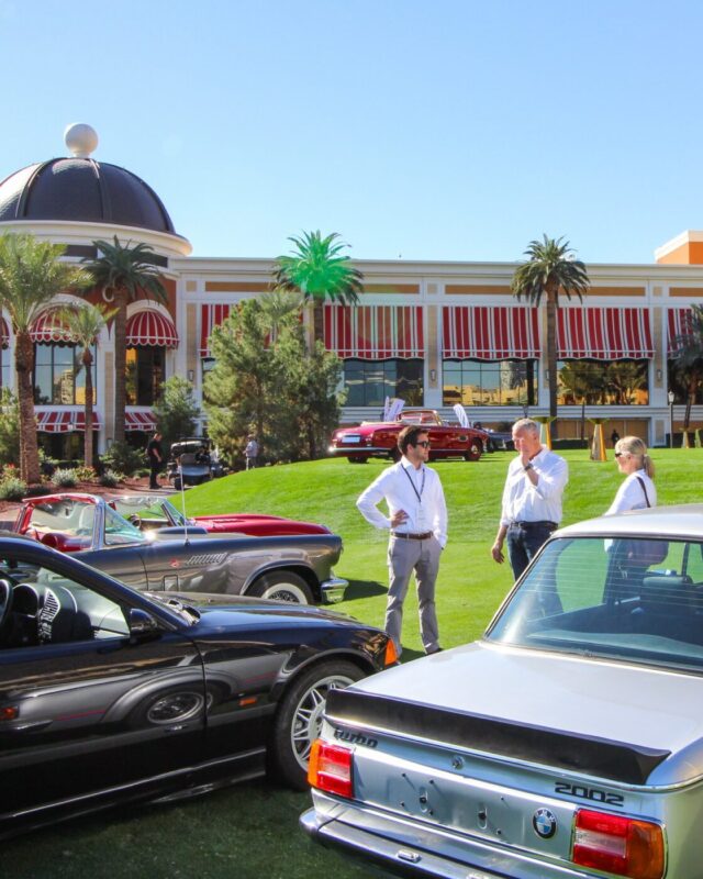 Three people stand and talk on a grassy lawn surrounded by classic BMW cars at the Broad Arrow Las Vegas Auction, with a striped-awning building and palm trees in the background.