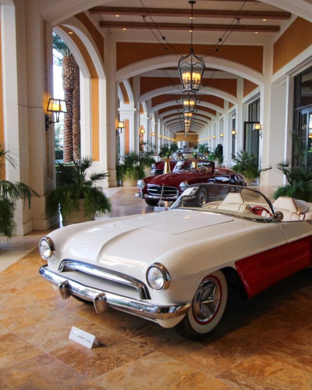 A white and red vintage convertible is displayed indoors on a tiled floor, with two other classic cars lined up behind it in a corridor with arches and hanging lights at the Broad Arrow Las Vegas Auction.