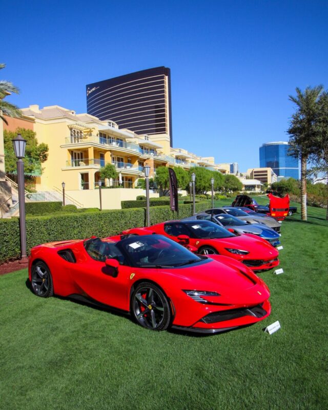 Four luxury sports cars, including two red Ferraris and a stunning Bugatti Veyron, are displayed on a well-kept lawn with hotels and palm trees in the background at the Broad Arrow Las Vegas Auction on a sunny day.