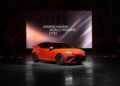 An orange Genesis GV60 Magma car is displayed in front of a large screen reading "Genesis Magma World Premiere" in a dark indoor venue with red accent lighting, showcasing the latest production model.