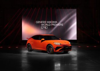 An orange Genesis GV60 Magma car is displayed in front of a large screen reading "Genesis Magma World Premiere" in a dark indoor venue with red accent lighting, showcasing the latest production model.