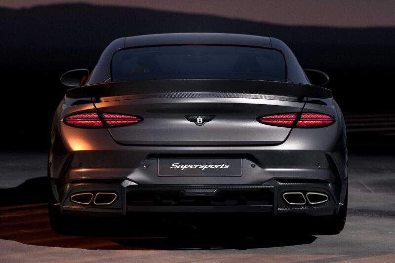 Rear view of a dark gray Bentley Supersports, a driver-focused Continental GT with illuminated tail lights, quad exhausts, and a prominent rear spoiler, parked on a road at dusk.
