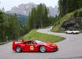 A red Ferrari sports car and a white classic convertible drive on a winding mountain road surrounded by trees and rocky hills.