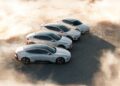 Four white electric cars are parked in a staggered formation on a sandy surface with dust and sunlight surrounding them.