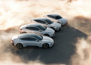 Four white electric cars are parked in a staggered formation on a sandy surface with dust and sunlight surrounding them.