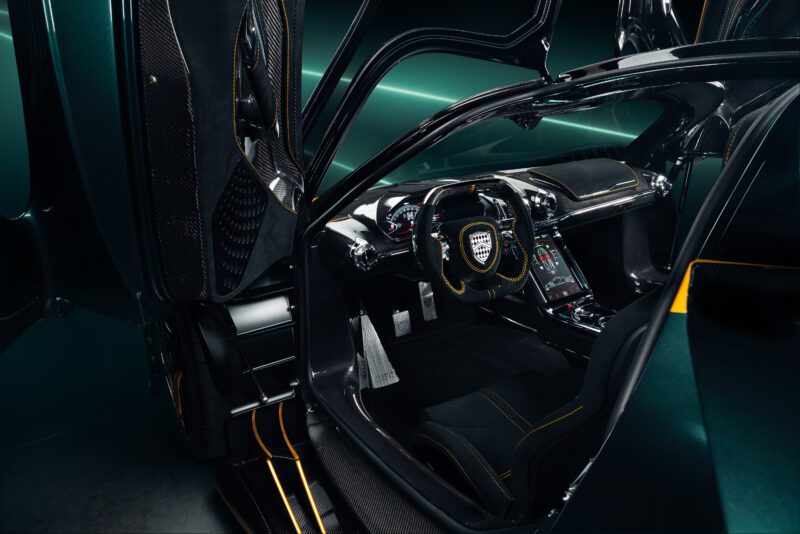 View of a luxury Tuatara Aggressor hypercar interior with butterfly doors open, showing the steering wheel, digital display, and carbon fiber details with yellow accents.