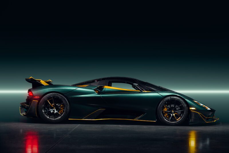 A low-angle side view of the Tuatara Aggressor, a sleek, dark green SSC North America hypercar with yellow accents, parked indoors on a reflective surface against a gradient backdrop.