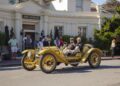 Two men drive a vintage yellow open-top car past a group of people outside a building labeled "The Lodge," capturing the spirit of automotive artistry that's defined 75 years of the Pebble Beach Concours d’Elegance.