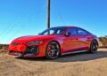 A red 2025 Audi RS e-tron GT Performance electric sports car is parked on a dirt road under a clear blue sky, with utility poles and wires in the background.
